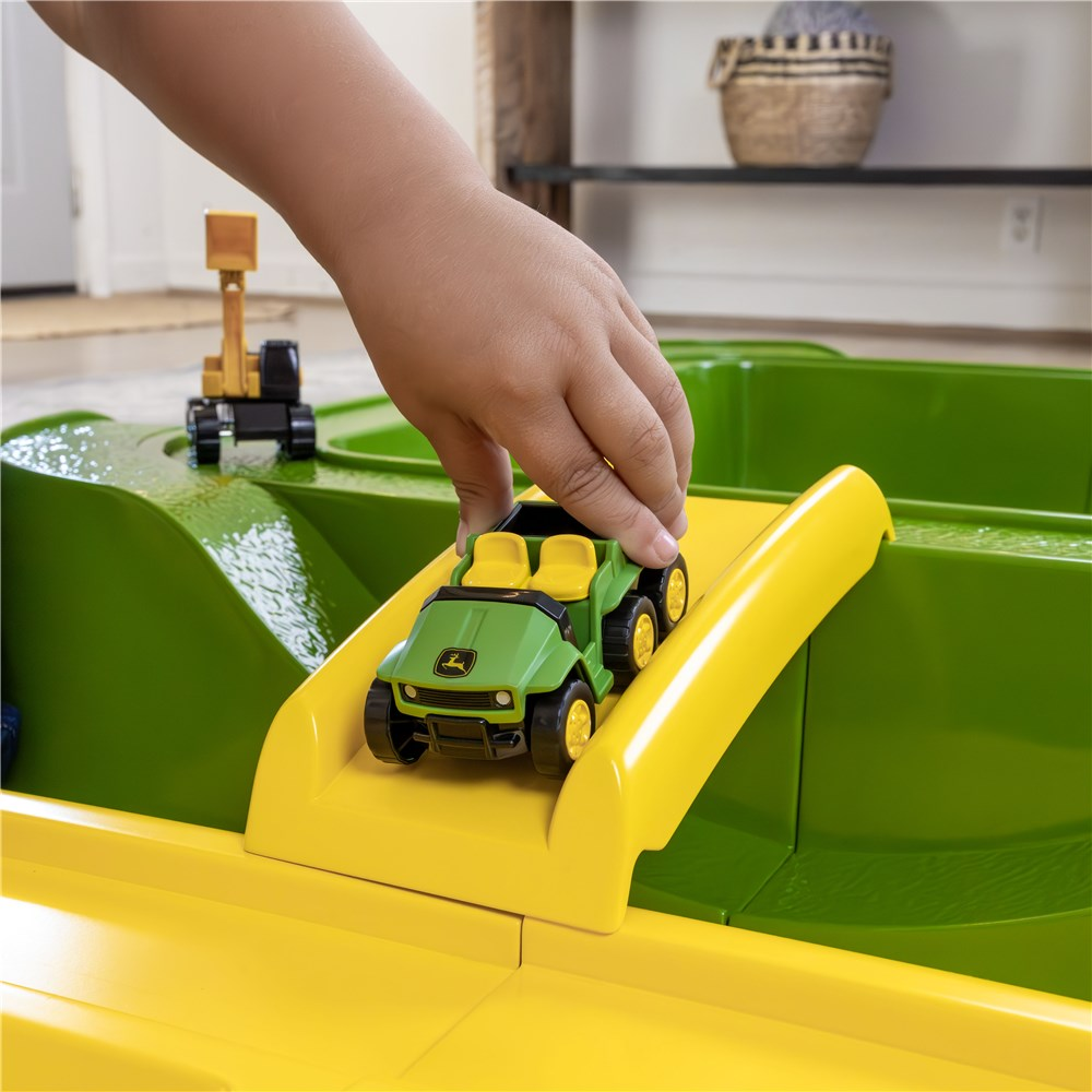 Child guiding a green John Deere toy vehicle down the yellow ramp built into the sand pit